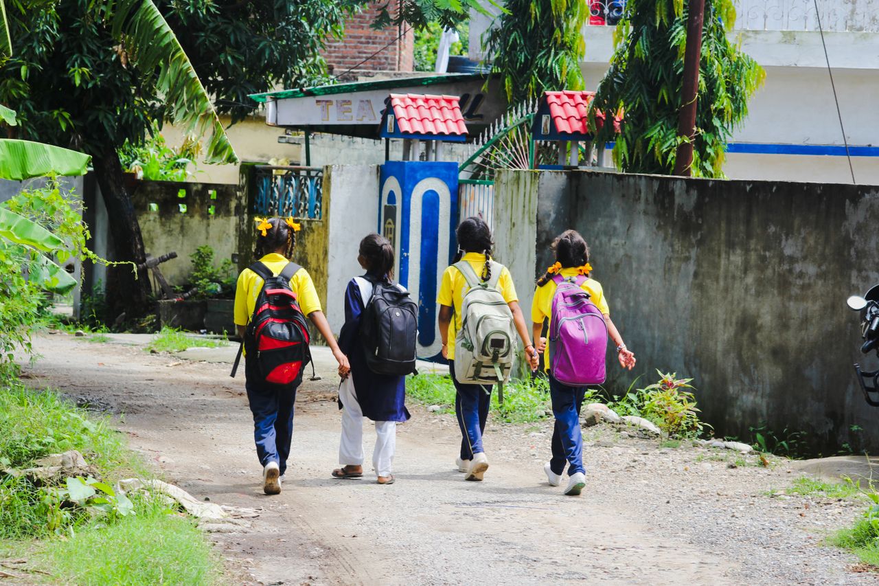 Four school children walk on an unpaved road, carrying backpacks, surrounded by greenery.